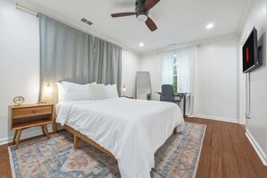 Habitación de hotel con cama, escritorio, silla, televisor, ventilador de techo y ventana con cortinas.