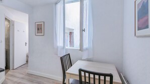 Ein Zimmer mit einem weißen Tisch, einem schwarzen Stuhl, einem Fenster mit weißen Vorhängen und einem gerahmten Bild an der Wand.