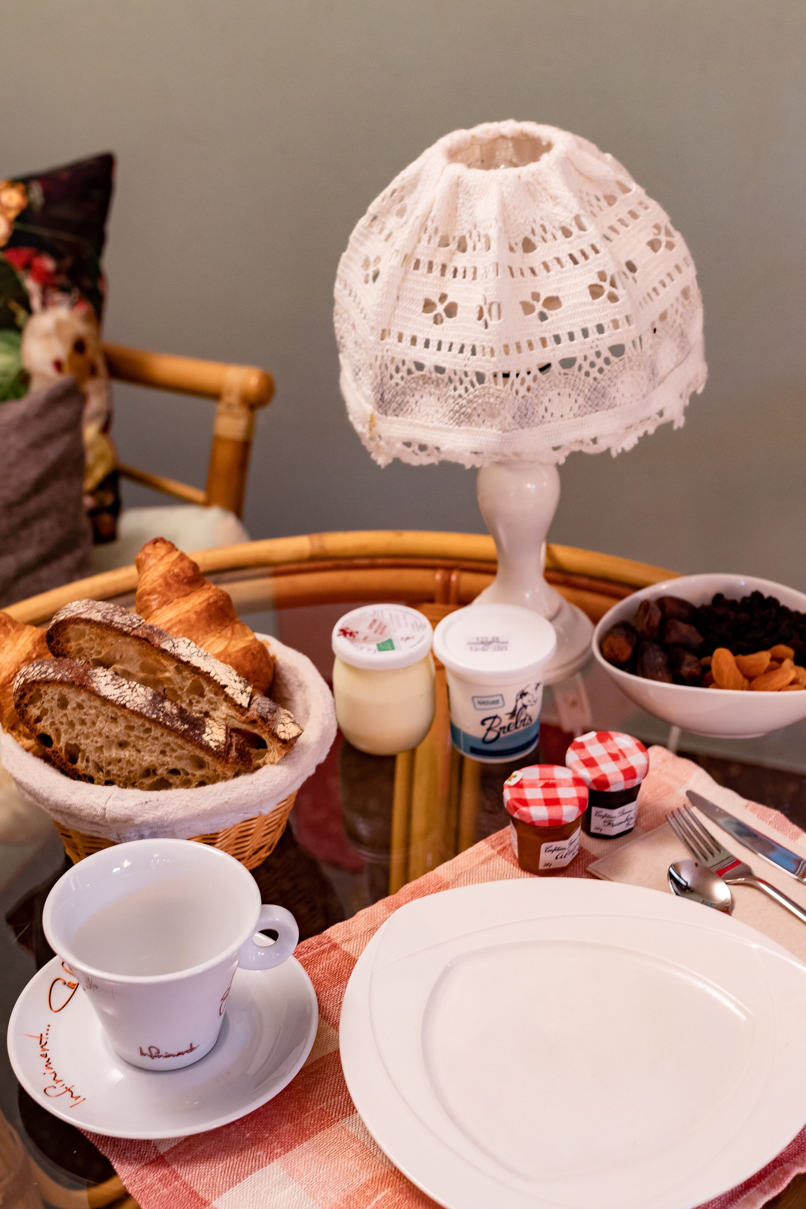 A table with a basket of croissants, a cup of coffee, a bowl of nuts, and a lace table lamp.