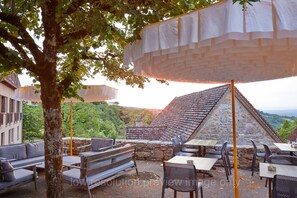 Un espacio al aire libre con mesas y bancos, rodeado de muros de piedra y un árbol.