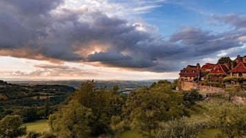 A hillside view of a traditional house with a stone foundation, surrounded by lush greenery and a dramatic sky.