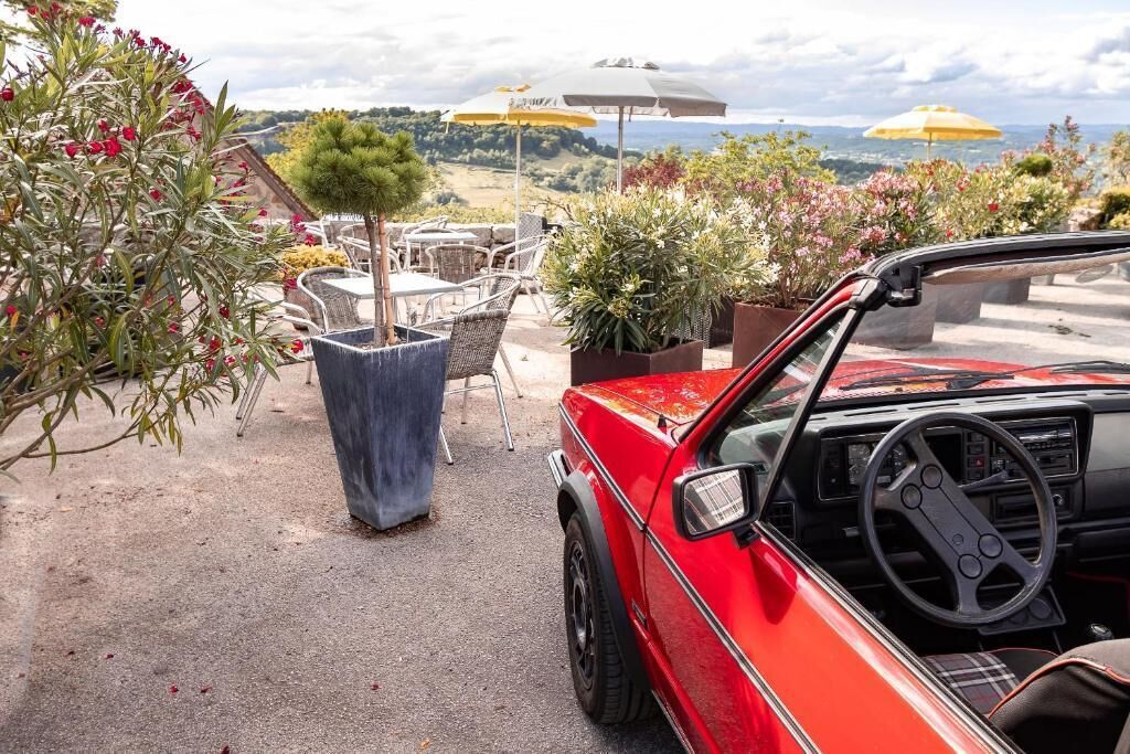 A red convertible parked on a patio with outdoor furniture, surrounded by potted plants and umbrellas.