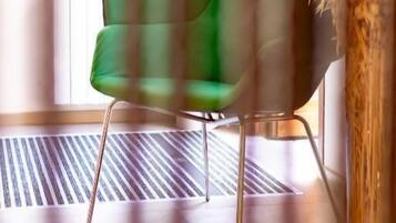 A green upholstered chair with metal legs in a room with a radiator and a tall vase with dried plants.