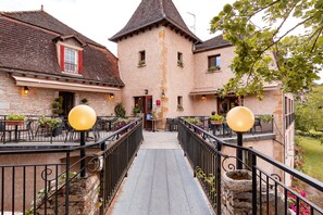 Un edificio de piedra con una entrada central, zona de estar al aire libre con mesas y sillas, y un pequeño puente que lleva a la entrada.