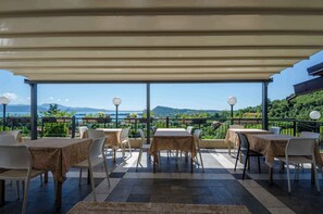 An outdoor patio with tables and chairs, a view of the sea, and mountains in the background.
