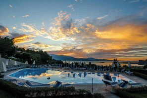 A swimming pool with lounge chairs and umbrellas, surrounded by greenery and a scenic sunset view.