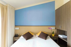 A hotel room with a large bed, a nightstand with a phone, a small table with a plant, and a wardrobe.