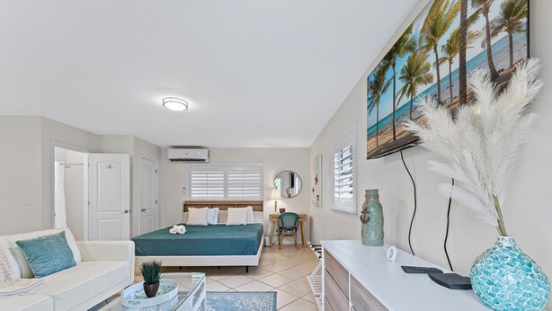 Un dormitorio bien iluminado con una cama, un escritorio y una silla. Cuenta con un ventanal con persianas, un aparador blanco y un cuadro de palmeras en la pared.