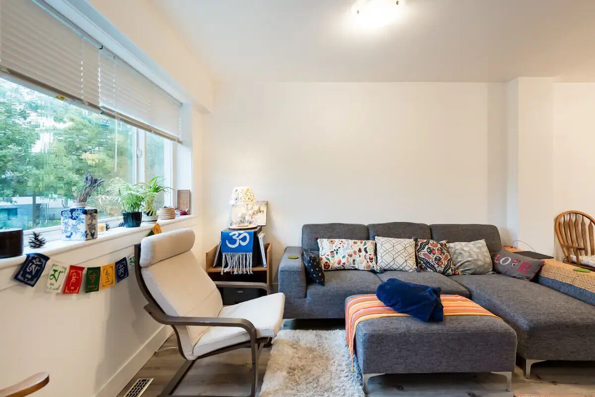 Een moderne woonkamer met een grijze bank, een witte fauteuil en een kleine tafel met een lamp. Er staan planten op de vensterbank en een reeks kleurrijke vlaggetjes.