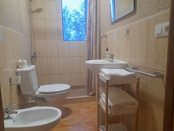 A small bathroom with a toilet, bidet, sink, and a window with a view of trees. - EntreOlivos (Badajoz)