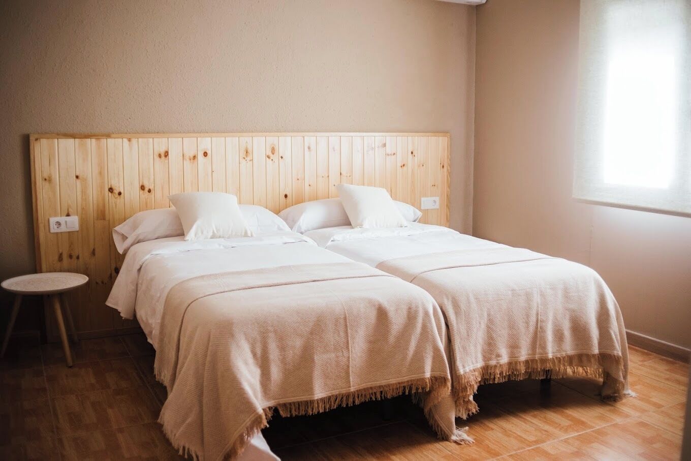 A double bed with a wooden headboard, a round stool, and a window with blinds.