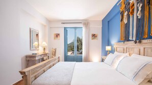 Ein Hotelzimmer mit einem großen Bett, einem Holz-Kopfteil, einem Schreibtisch mit Lampe und Blick ins Freie durch eine blaue Tür.