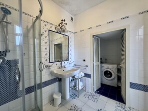 A bathroom with a glass-enclosed shower, a sink with a mirror, a washing machine, and patterned tiles. - YourHome - Assia House Praiano (Praiano)