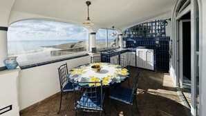 A sunlit patio with a table and chairs, a view of the sea, and a kitchen area with blue cabinets. - YourHome - Assia House Praiano (Praiano)