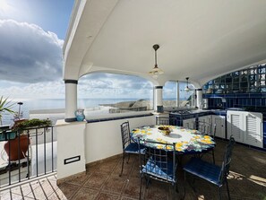 A sunlit patio with a round table and chairs, a view of the sea, and a kitchen area with blue tiles. - YourHome - Assia House Praiano (Praiano)
