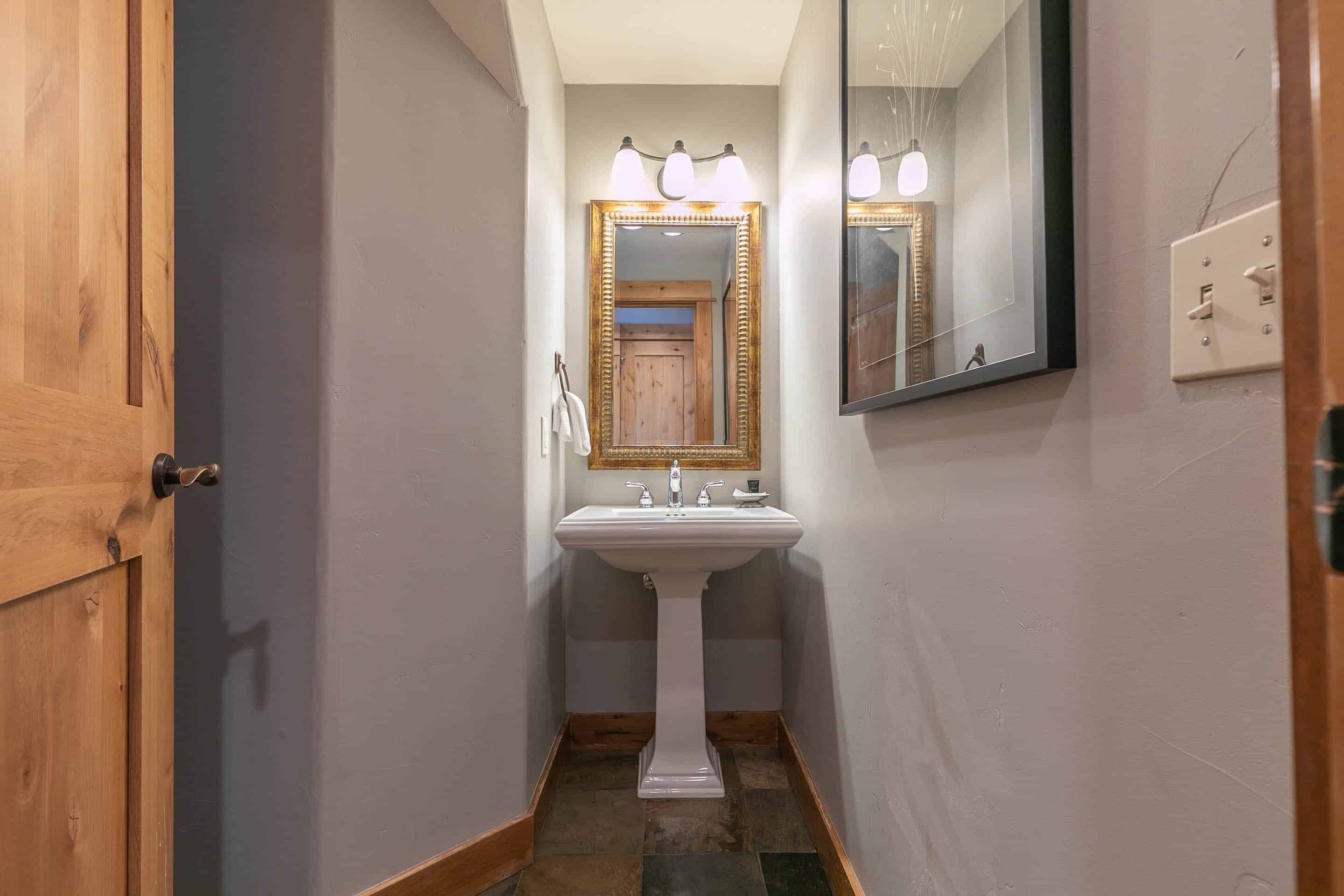 A narrow hallway with a white pedestal sink, wooden door, and a mirror with lights above it.