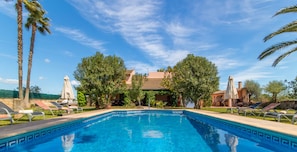 A clear blue swimming pool with lounge chairs and umbrellas, surrounded by trees and a building with a tiled roof.