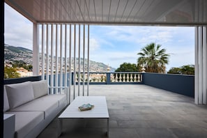 A balcony with a white sofa, a small table, and a view of a cityscape with mountains and palm trees. - Luna Rossa - Artcooking by Homie (Funchal)