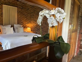 A hotel room with a brick wall, a wooden bed frame, white bedding, and a large potted plant with white flowers.