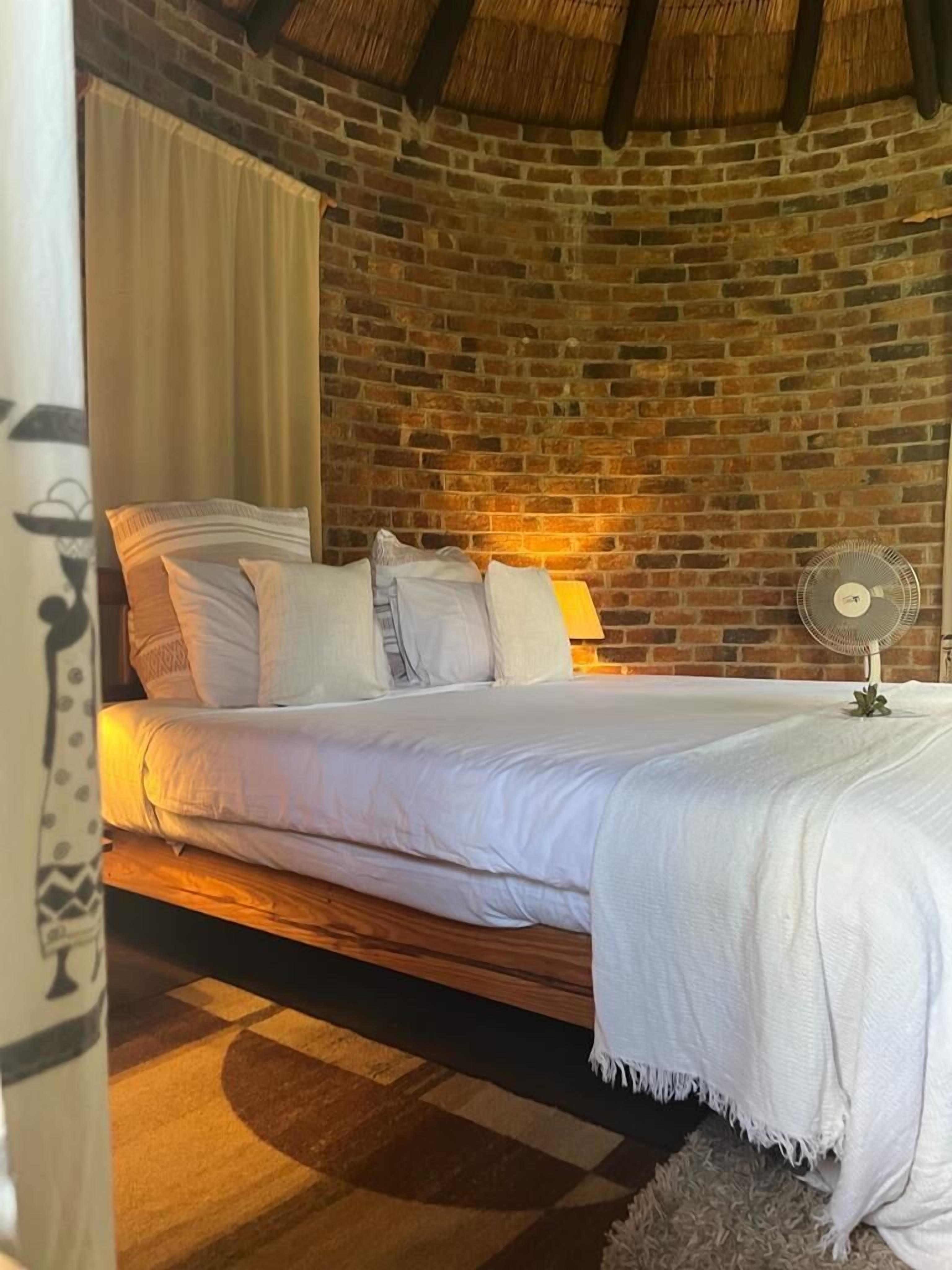 A room with a brick wall, a bed with white bedding, a small fan, and a curtain.