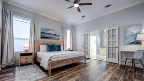 A bedroom with a wooden bed, a desk, and a chair. There is a window with curtains, a ceiling fan, and a painting on the wall. - Lei'd Back * Golf Cart Included & Pet Friendly * Salt Water Vacations (Destin)