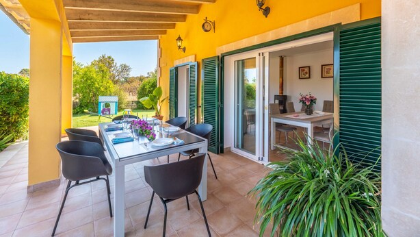 Un patio al aire libre con una mesa y sillas, un espacio cubierto con techo de madera y vistas a un jardín.