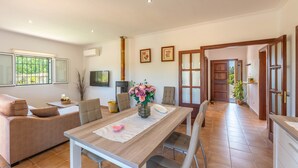 Una sala de estar moderna con un sofá, una mesa de comedor y sillas. Hay un televisor montado en la pared, una chimenea y un ventanal con vistas a la vegetación.