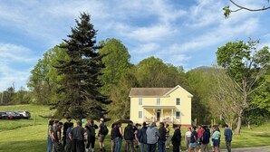 A group of people standing in front of a yellow house with a porch, surrounded by trees and a gravel path. - Peaceful Farmhouse w/ Fire Pit & Updated Comforts (Monroe)