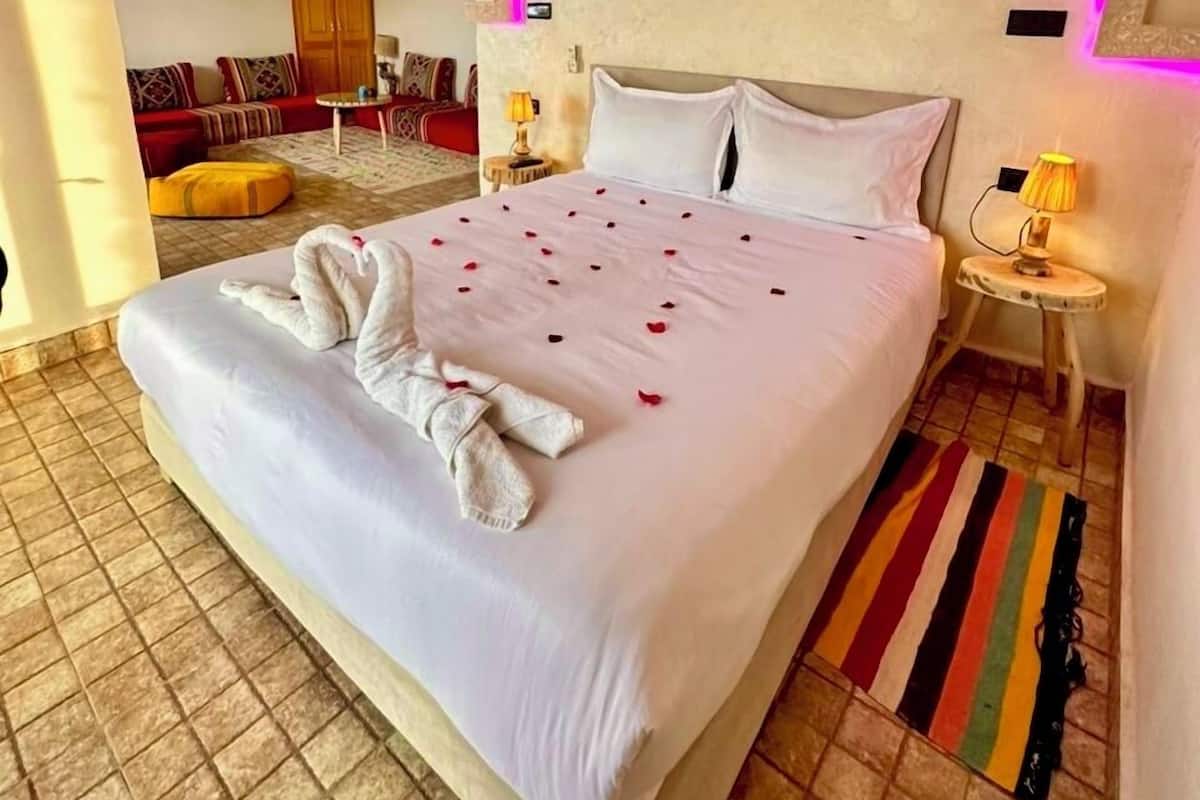A neatly made bed with a swan-shaped towel and rose petals, a round mirror, and a small table with a lamp.