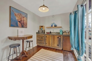Une cuisine avec des meubles de rangement en bois, un tabouret de bar, un luminaire suspendu et une fenêtre avec des rideaux.