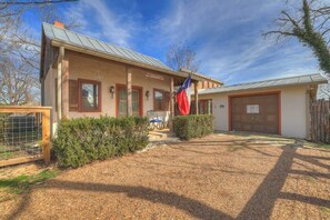 A two-story building with a metal roof, a flagpole with a flag, a covered garage, and a gravel driveway. - TX 2-Step Guesthouse:Pet Friendly-3Min to Downtown (Fredericksburg)