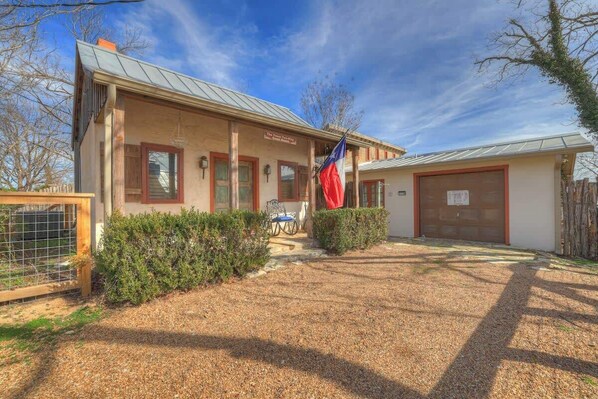A two-story building with a metal roof, a flagpole with a flag, a covered garage, and a gravel driveway.