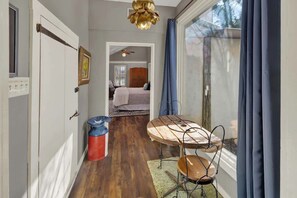 A narrow hallway with a wooden floor, a table with chairs, a red trash bin, and a window with blue curtains.