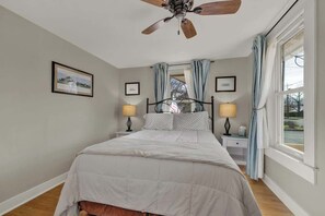 Un dormitorio con cama, dos mesitas de noche con lámparas, ventilador de techo y ventana con cortinas.