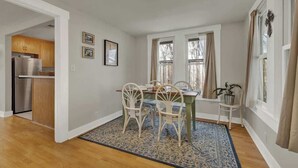 A dining area with a wooden table, white chairs, and a patterned rug. There are two windows with curtains, a kitchen visible through an open door, and a small plant on a side table.