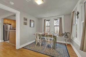 A dining area with a wooden table, white chairs, and a patterned rug. There are two windows with curtains, a kitchen visible through an open door, and a small plant on a side table.