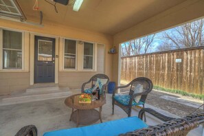 A covered patio with wicker furniture, a small table with a bottle and glasses, and a view of a wooden fence.
