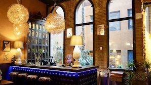 A bar area with a cushioned seating arrangement, hanging pendant lights, and a view through large arched windows. - Hotel Lincoln Lounge (Barcelona)