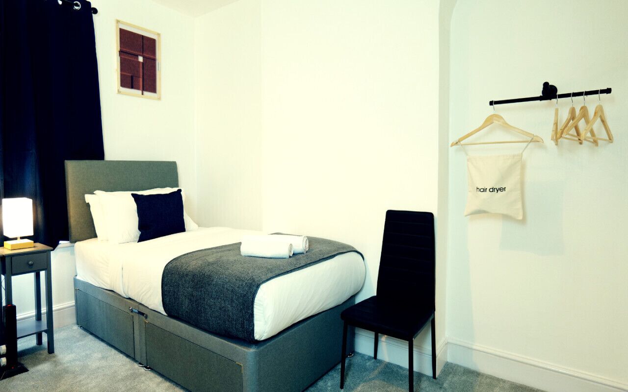 Un dormitorio con una cama, una silla, una mesita de noche, un perchero y una ventana pequeña.