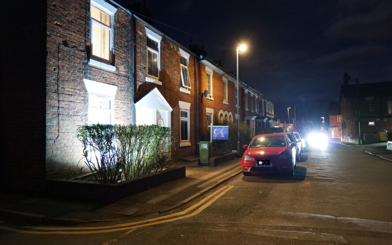 A residential street at night with houses, a red car parked on the side, and street lighting.