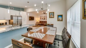 Una cocina moderna con una isla central, electrodomésticos integrados, iluminación colgante y un comedor con una mesa grande y asientos acolchados.