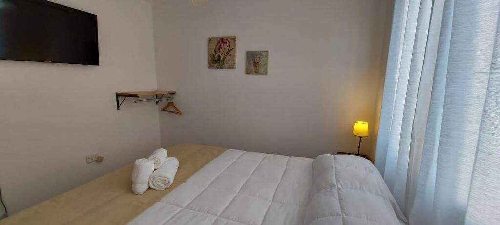 Habitación de hotel con cama, televisor montado en la pared, mesita de noche con lámpara y ventana con cortinas.