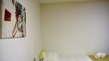 A single bed with a white duvet, a bedside lamp, and a rolled towel on the floor.