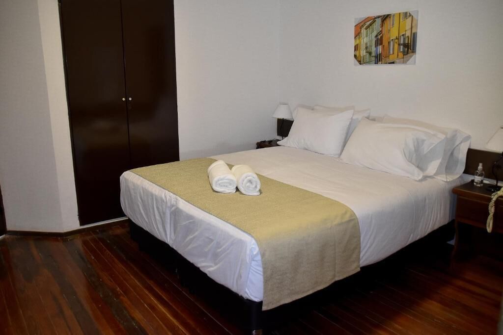 A neatly made bed with white linens and a beige throw, a nightstand with a lamp, and a painting of buildings on the wall.
