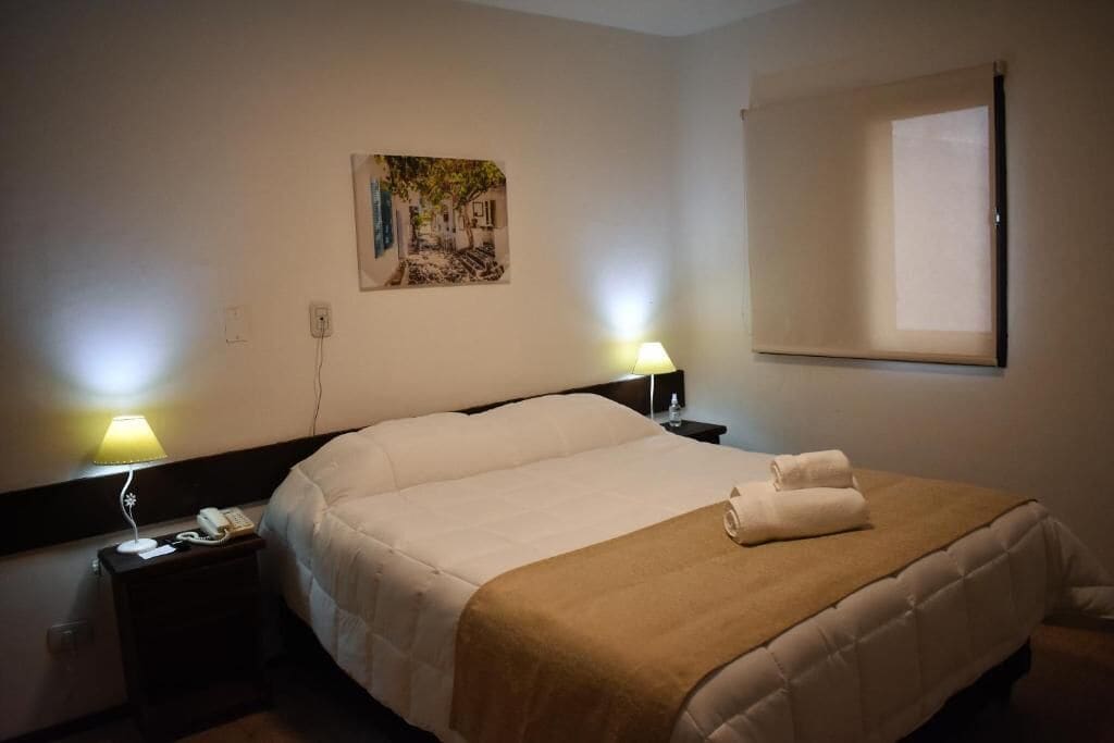 Hotelkamer met een bed, nachtkastje met lamp, schilderij aan de muur en raam met jaloezieën.