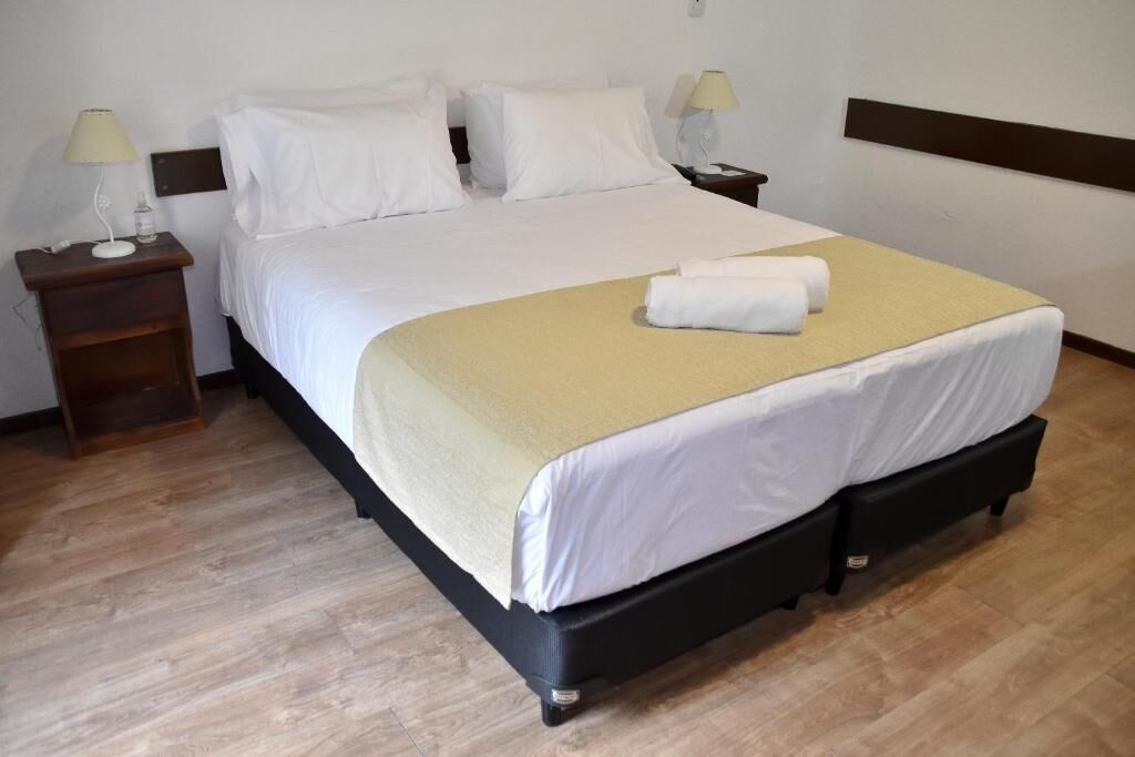 Una cama bien tendida con sábanas blancas y un plaid color beige, una mesita de noche con una lámpara y otra mesita con una lámpara.