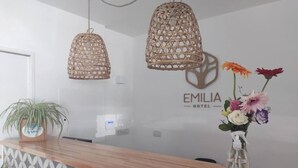 A hotel reception area with a wooden counter, a potted plant, and a wall-mounted logo for Emilia Hotel. - EMILIA HOTEL (Rosario)