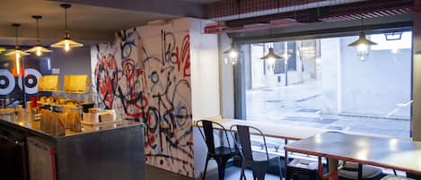 Interior moderno de una cafetería con arte callejero en la pared, un mostrador con productos alimenticios y vistas a la calle a través de grandes ventanales.