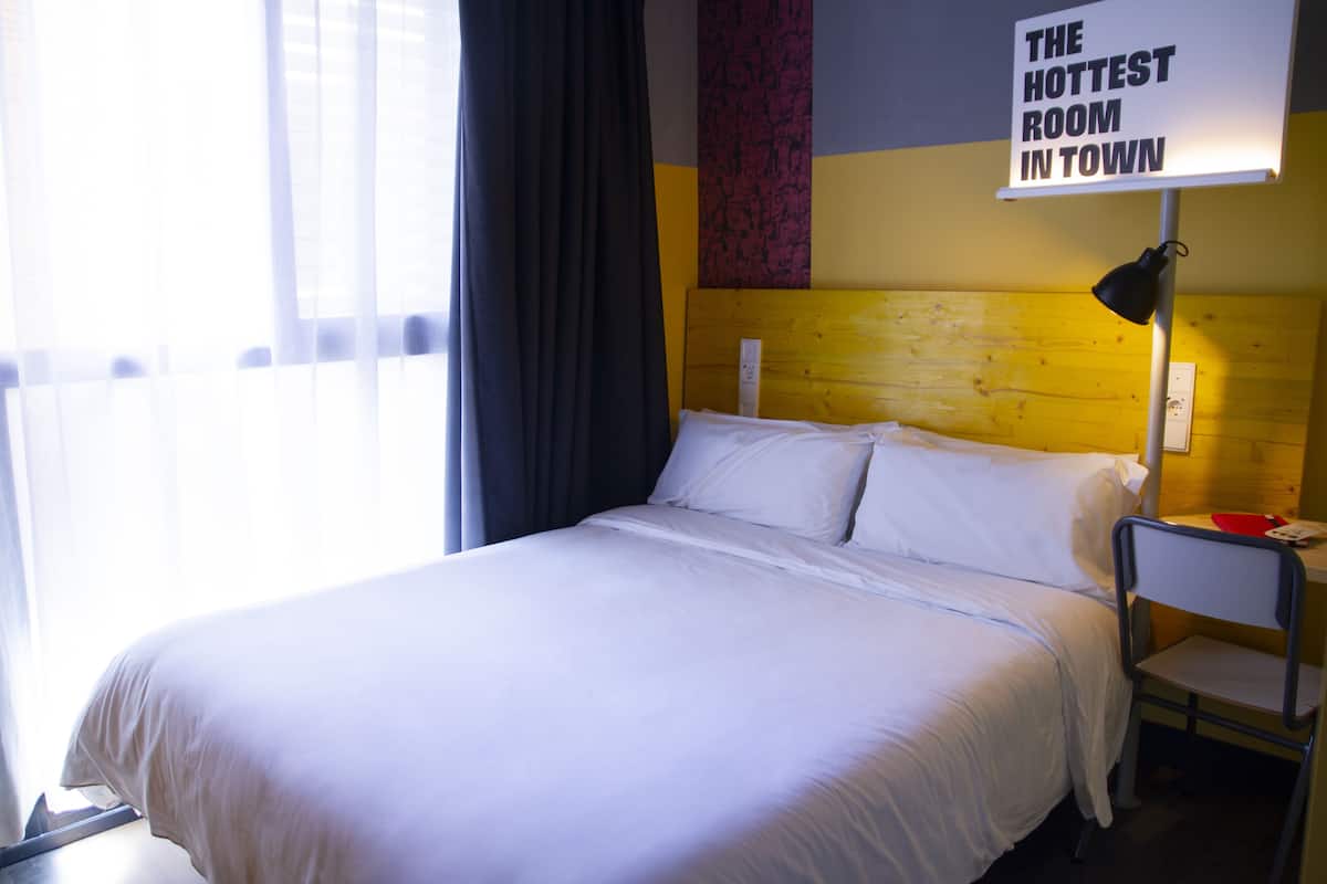 Quarto de hotel com uma cama, mesa de cabeceira, luminária e um letreiro com a inscrição 'THE HOTTEST ROOM IN TOWN'.