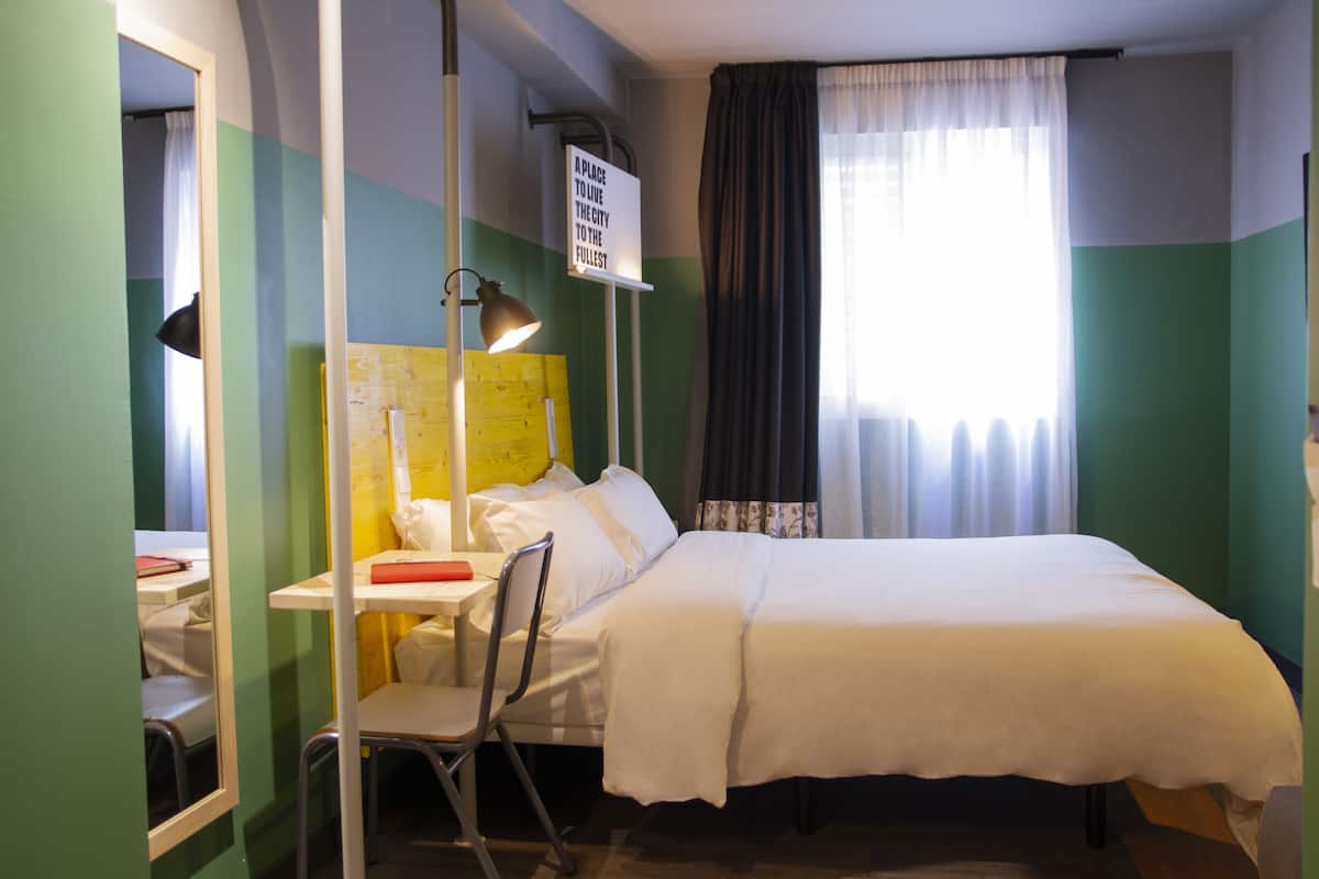 Quarto de hotel com cama, escrivaninha com cadeira, abajur e um aviso na parede.